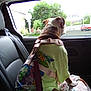 animal_print, backseat, blanket, brown_and_white_dog, car_interior, daylight, dog, harness, leash, leather_seat, looking_out_window, pet, road, sitting, street, trees, tshirt, urban, vehicle, window