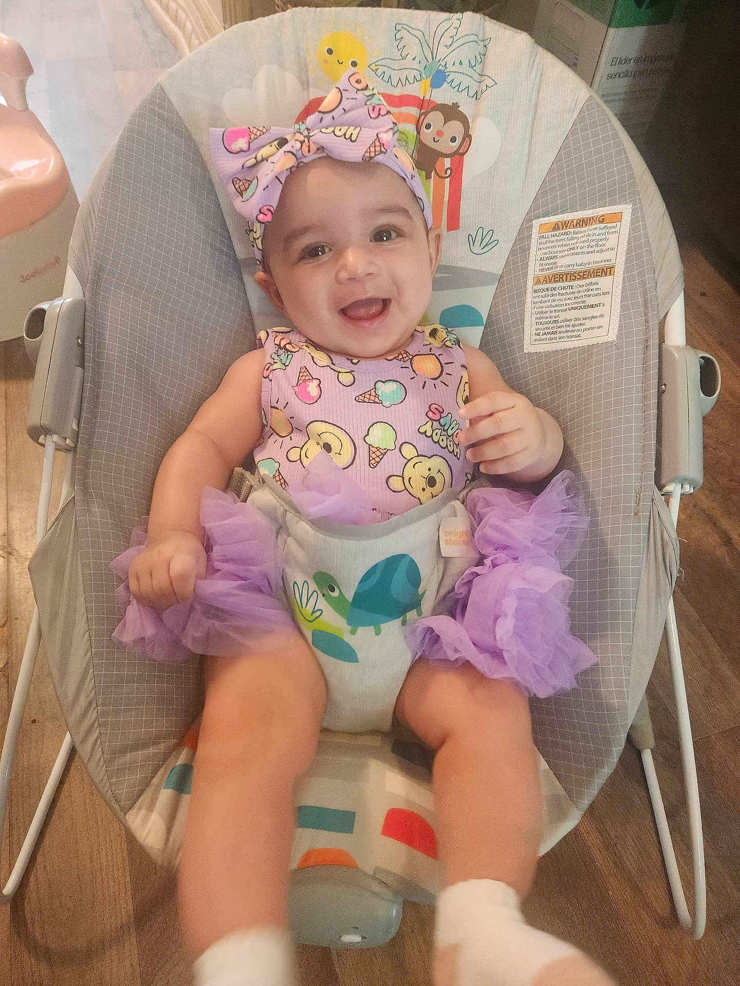 Trinity is registered to the contest to win money with this photo: baby, infant, smiling, child, bouncer, purple_clothing, headband, tutu, indoor, wooden_floor, cute, happy, person, face, seat, playful, animal_print, toddler, sitting, joyful