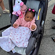 Annalise is registered to the contest to win money with this photo: baby, stroller, blanket, toy, unicorn, pink_jacket, hair_ties, outdoor, pavement, child, infant, cute, person, seat, accessory, footwear, clothing, playful, bundle, curious