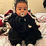 baby, child, pacifier, black_outfit, curly_hair, bedspread, blanket, socks, indoors, cute, portrait, seated, person, infant, soft_texture, warm_clothing, cozy, looking_at_camera, expression, toddler