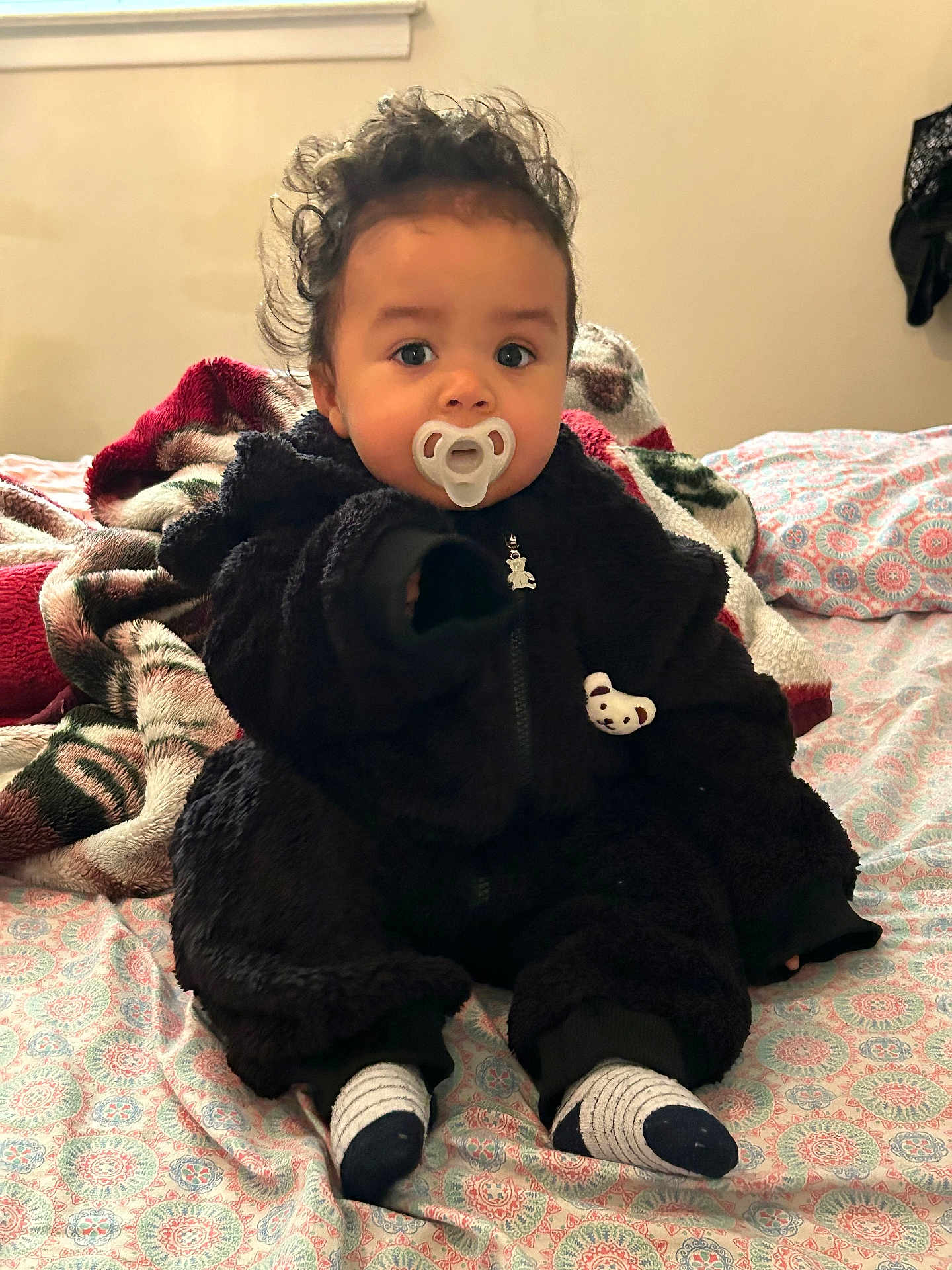 Josiah is registered to the contest to win money with this photo: baby, child, pacifier, black_outfit, curly_hair, bedspread, blanket, socks, indoors, cute, portrait, seated, person, infant, soft_texture, warm_clothing, cozy, looking_at_camera, expression, toddler