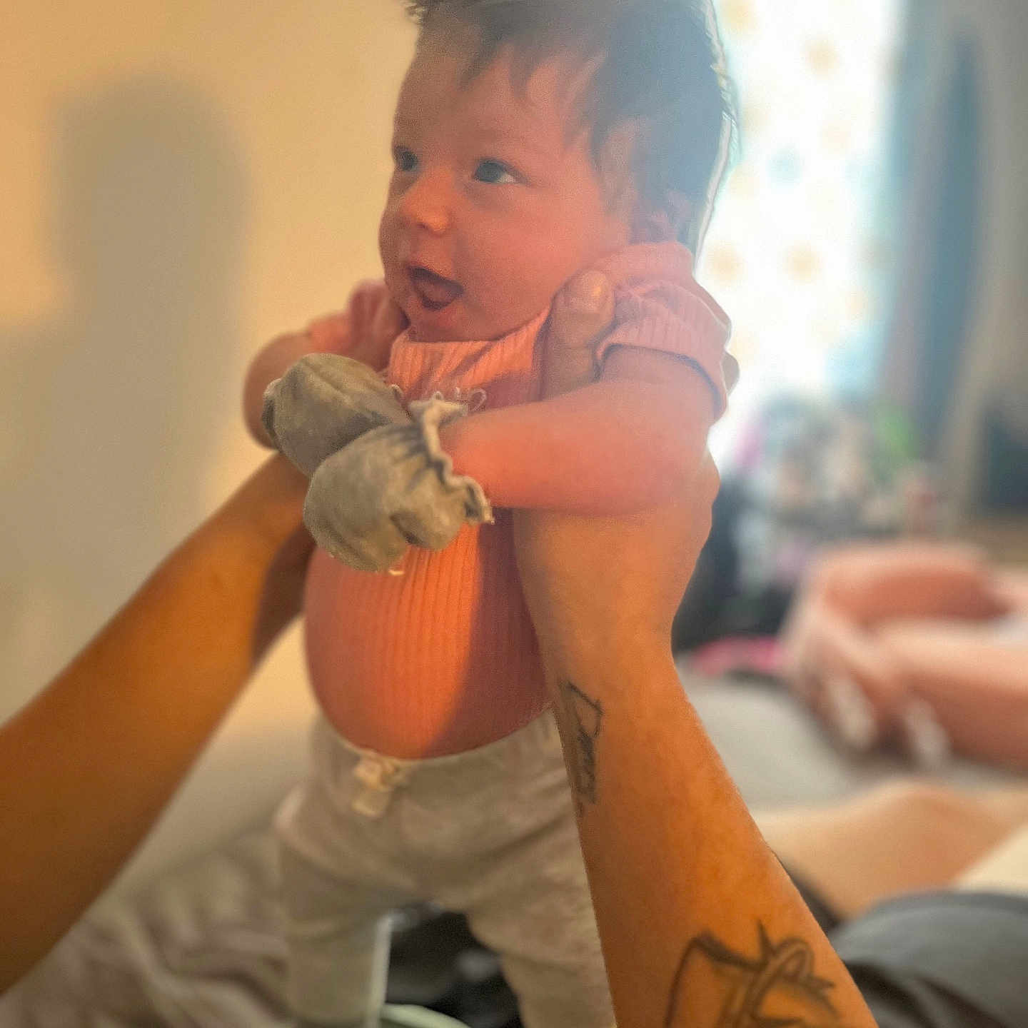 Joselynn joined the competition — help win amazing prizes! baby, blurred_background, child, cozy, curious_expression, cute, hands, holding, home, human, indoor, infant, mittens, person, pink_clothing, portrait, smiling, soft_lighting, tattoo, young