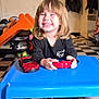 black_shirt, blonde_hair, blue_table, book, cartoon, child, coat, floor_tiles, furniture, happy, indoor, person, playing, red_toy_car, room, smiling, table, television, toy_controller, young
