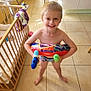 child, boy, smiling, indoor, tile_floor, swim_trunks, water_toys, inflatable_ring, crib, blanket, unicorn, kitchen, cabinet, sunlight, playful, barefoot, happy, young, person, home