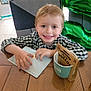 child, boy, smiling, envelope, table, chair, balloon, number_two, birthday, cup, package, indoor, wooden_table, plaid_shirt, happy, face, hands, celebration, gift, seated