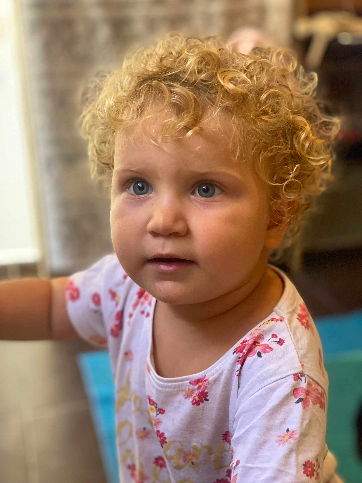 Emelin participe au concours pour gagner de l'argent avec cette photo : child, curly_hair, blonde_hair, blue_eyes, portrait, indoor, toddler, cute, face, person, clothing, white_shirt, floral_pattern, expression, looking_away, soft_background, skin, head, young, innocent