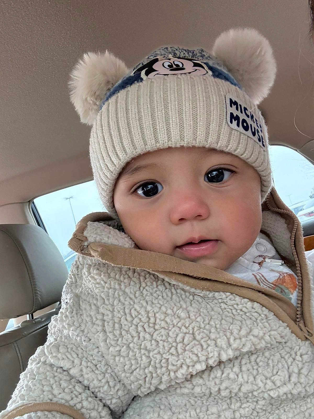 John is registered to the contest to win money with this photo: baby, child, hat, mickey_mouse, pom_poms, jacket, sherpa, cozy, face, eyes, car_interior, seat, clothing, winter_wear, cute, portrait, young_child, warm, close_up, infant