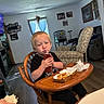 child, toddler, young_person, sitting, high_chair, juice_box, straw, drinking, food, fried_food, meal, table, wooden_furniture, living_room, armchair, couch, window, curtains, family_home, portrait