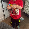 child, boy, toddler, blond_hair, standing, portrait, indoor, home, toy, beads, colorful, red_shirt, sportswear, black_pants, socks, pet_cage, cage, sofa, wooden_floor, playing