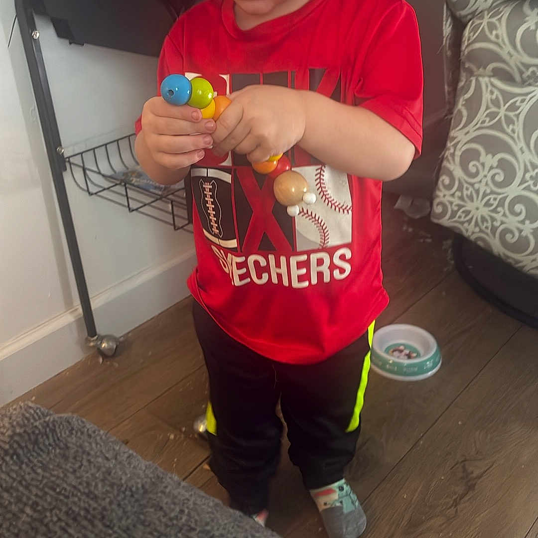 Hayse is registered to the contest to win money with this photo: child, boy, toddler, blond_hair, standing, portrait, indoor, home, toy, beads, colorful, red_shirt, sportswear, black_pants, socks, pet_cage, cage, sofa, wooden_floor, playing