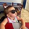 Athena joined the competition — help win amazing prizes! toddler, child, sunglasses, bib, table, menu, chair, indoor, restaurant, wooden_chair, tile_floor, person, young_child, casual, seated, hand, face, hair_bow, pink, clothing