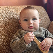 Andrea participe au concours pour gagner de l'argent avec cette photo : baby, child, blue_eyes, sweater, wooden_brush, couch, indoor, cozy, curious_expression, knitted_clothing, seated, soft_lighting, portrait, infant, cute, handheld_object, comfortable, home, casual_clothing, young_child