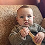 baby, child, blue_eyes, sweater, wooden_brush, couch, indoor, cozy, curious_expression, knitted_clothing, seated, soft_lighting, portrait, infant, cute, handheld_object, comfortable, home, casual_clothing, young_child