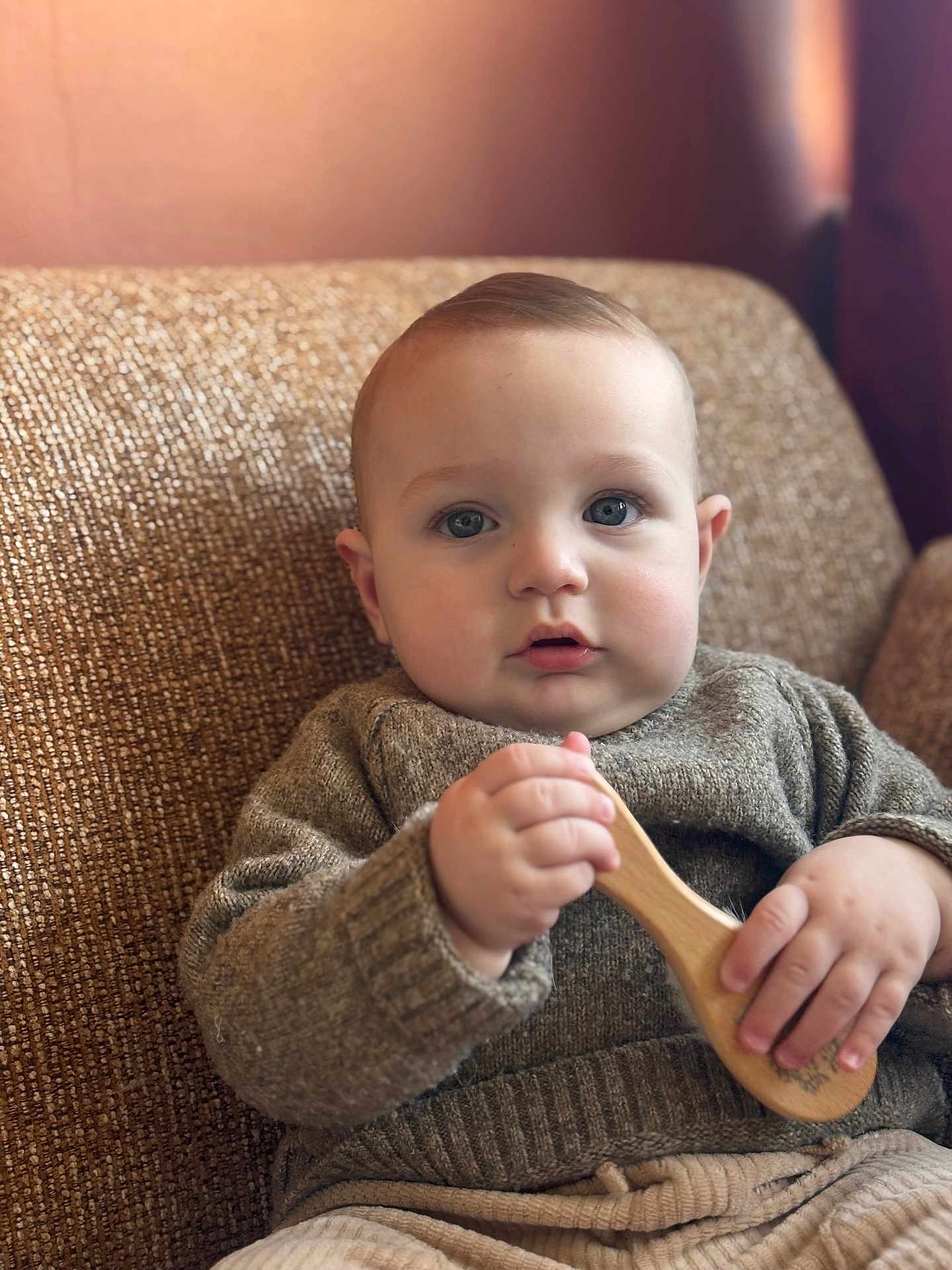 Andrea participe au concours pour gagner de l'argent avec cette photo : baby, child, blue_eyes, sweater, wooden_brush, couch, indoor, cozy, curious_expression, knitted_clothing, seated, soft_lighting, portrait, infant, cute, handheld_object, comfortable, home, casual_clothing, young_child