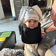 Liam is registered to the contest to win money with this photo: baby, bunny_hat, black_bib, tattooed_arm, indoor, person, book, baby_socks, table, couch, floor, window_light, cute, wide_eyes, holding, child, clothing, home, infant, portrait