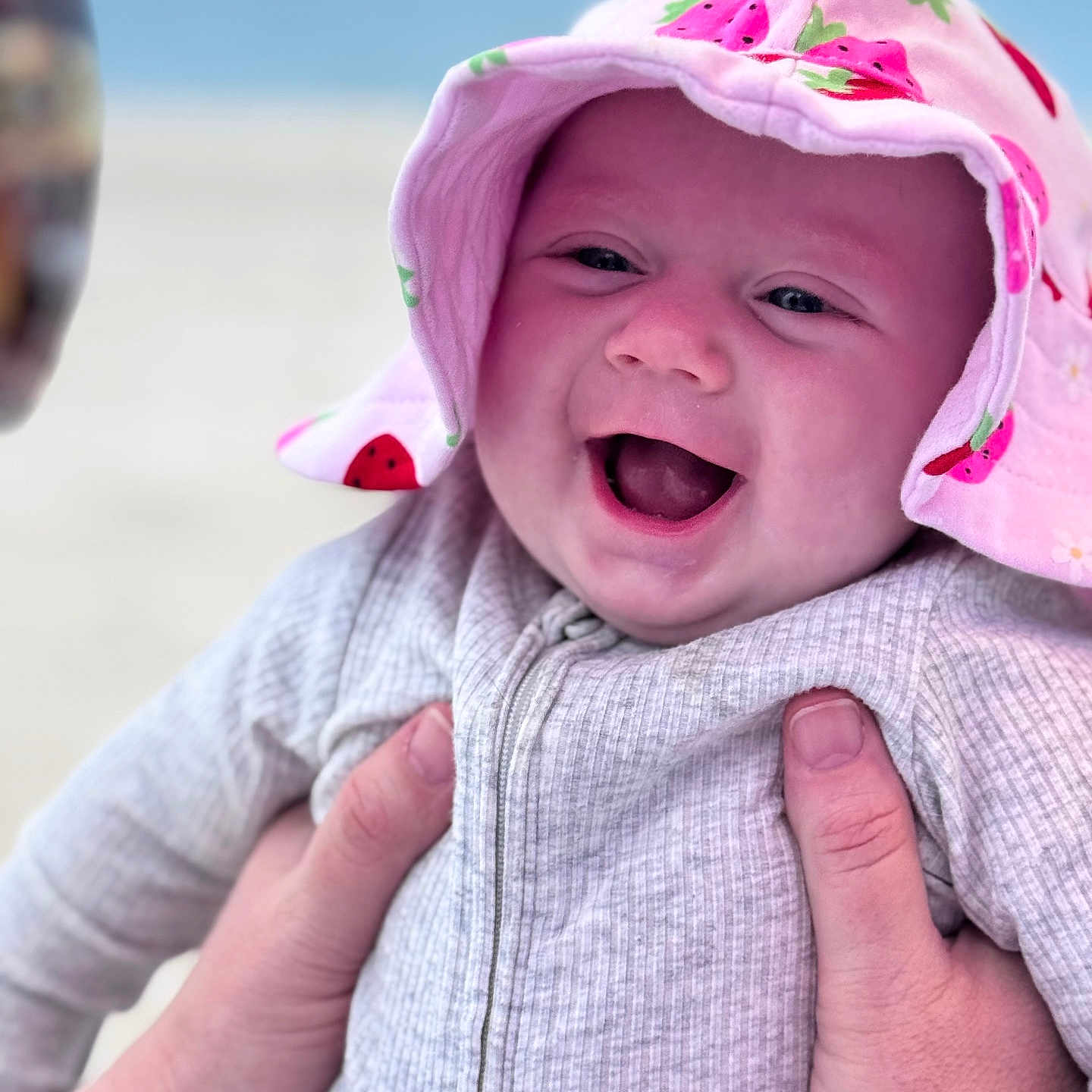 Willow joined the competition — help win amazing prizes! baby, beach, blurred_background, child, closeup, cute, gray_outfit, hands, happy, hat, infant, joyful, ocean, outdoor, person, pink_hat, portrait, smiling, strawberry_pattern, sunhat