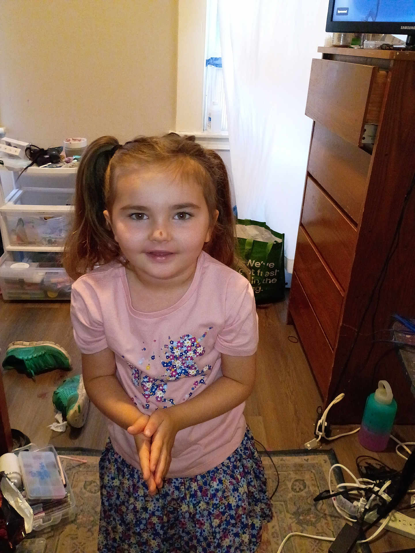 Rhea is registered to the contest to win money with this photo: child, girl, indoor, pigtails, pink_shirt, floral_skirt, smiling, standing, wooden_floor, dresser, shoes, plastic_storage, rug, curtain, window, water_bottle, power_strip, clutter, toy, person