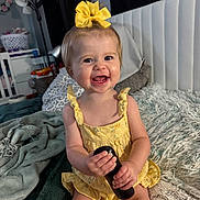 Adalynn is registered to the contest to win money with this photo: toddler, child, smiling, yellow_bow, yellow_dress, remote_control, bed, blanket, pillow, indoor, happy, person, furniture, head, face, skin, hair, sitting, cozy, room