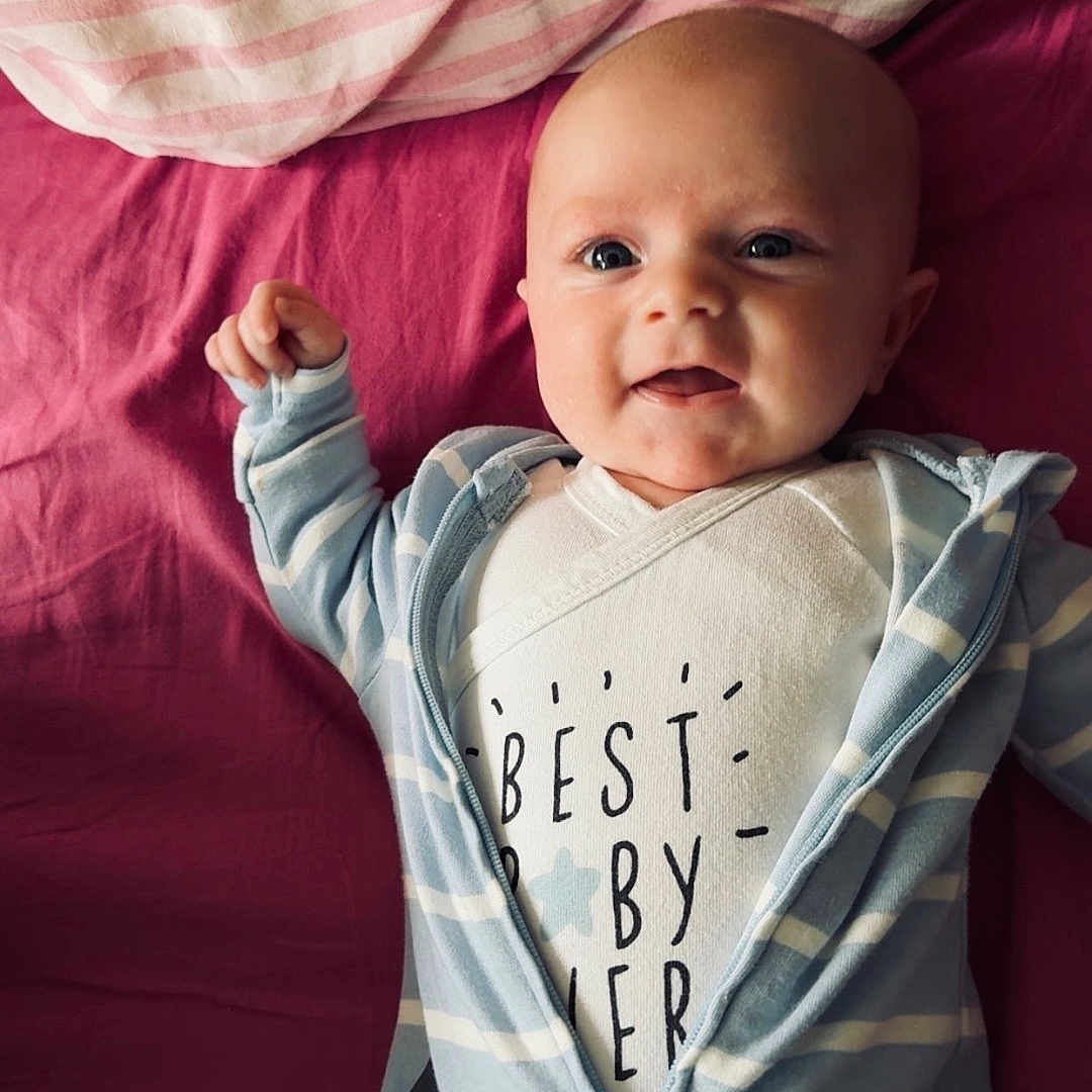 Noah participe au concours pour gagner de l'argent avec cette photo : baby, blue_onesie, child, closeup, cute, face, happy, indoor, infant, lying_down, newborn, person, pink_bed, portrait, raising_arm, smiling, soft_lighting, striped_pillow, text_on_clothing, white_shirt