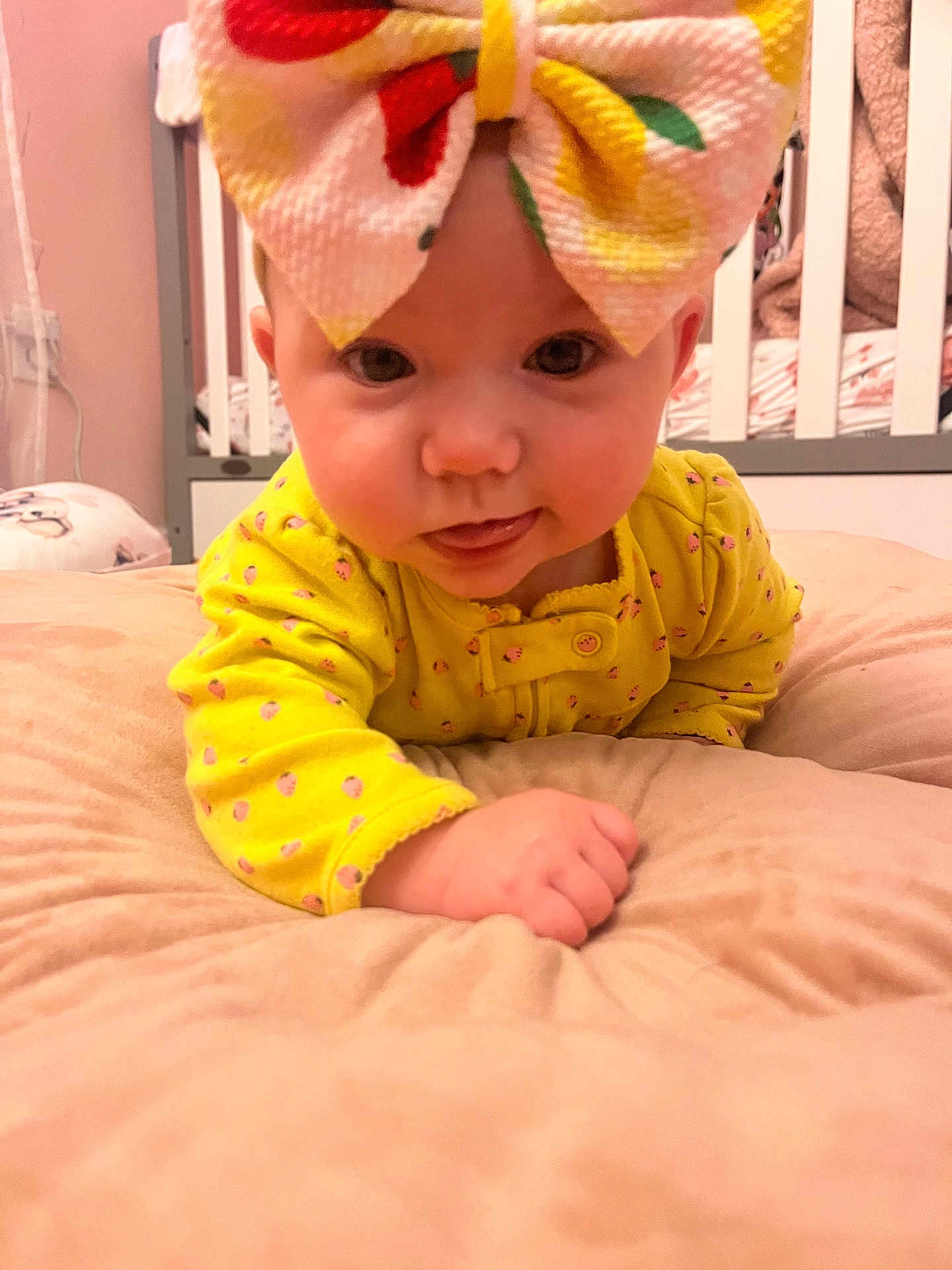 Isabella is registered to the contest to win money with this photo: baby, child, infant, yellow_clothing, headband, bow, tongue_out, cushion, crib, nursery, bedroom, soft_toys, cute, indoors, person, smiling, happy, face, lying_down, closeup