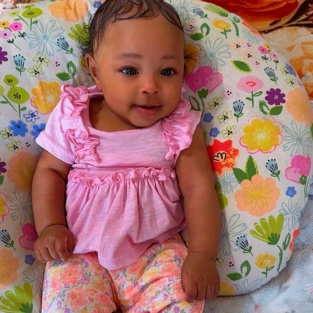Ahnarie is registered to the contest to win money with this photo: adorable, baby, bedspread, blanket, child, colorful, cozy, cute, floral_pattern, floral_pillow, hair_bow, hands, indoor, infant, patterned_pants, pink_clothing, portrait, sitting, smiling, socks