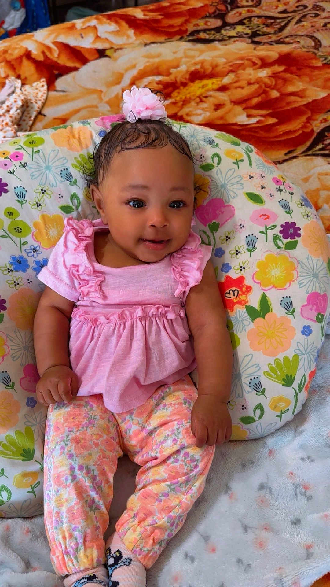 Ahnarie is registered to the contest to win money with this photo: baby, infant, child, pink_clothing, hair_bow, floral_pillow, floral_pattern, patterned_pants, blanket, bedspread, sitting, smiling, adorable, portrait, indoor, cozy, socks, hands, cute, colorful
