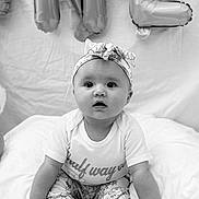 Emerson is registered to the contest to win money with this photo: baby, infant, headband, onesie, pants, floral_pattern, sitting, expression, balloons, celebration, portrait, black_and_white, cute, child, feet, hands, soft_surface, clothing, indoor, face