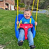 blue_shirt, blue_swing, brick_wall, child, daylight, footwear, fun, grass, greenery, happy, house, jeans, outdoor, play, red_safety_bar, smiling, swing, toddler, trees, yellow_ropes
