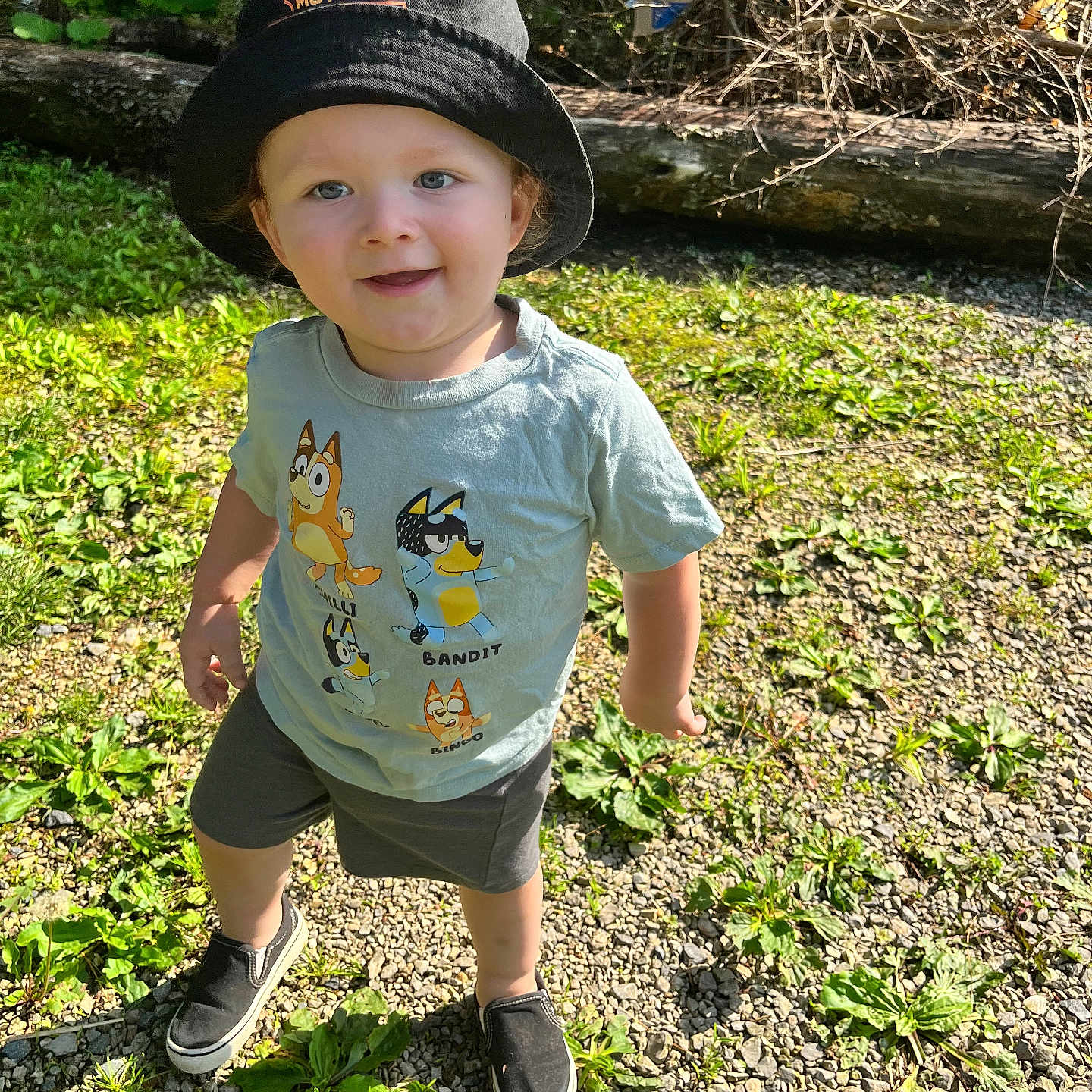 Brodhi is registered to the contest to win money with this photo: toddler, child, hat, bucket_hat, tshirt, cartoon, outdoor, sunny, gravel, plants, greenery, log, twigs, smiling, casual_clothing, footwear, shoes, nature, daylight, person