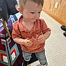casual_clothing, child, curious, curly_hair, flooring, footwear, home_interior, indoor, jeans, orange_shirt, person, rug, shoes, small_hands, smile, snack, standing, toddler, wood_paneling, young_child