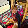 adult_legs, backward_cap, black_shoes, cap, chair, child, clothing, cushion, floor, footwear, geometric_pattern, home, indoor, nike, person, rug, sitting, smiling, sweatpants, toddler