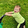 child, toddler, grass, green_shirt, black_shorts, toy, lawnmower, outdoor, playing, happy, smiling, grass_field, summer, fun, active, barefoot, sunlight, nature, cute, young