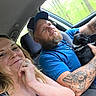 car, couple, woman, man, puppy, dog, tattoo, seatbelt, window, nature, trees, smile, cap, blue_shirt, driving, indoor, portrait, happy, pet, road_trip