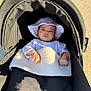baby, stroller, hat, blanket, outdoor, sunlight, infant, child, person, clothing, face, hand, headwear, baby_carriage, shadow, gravel, relaxing, cute, young_child, portrait