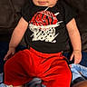 baby, child, infant, portrait, sitting, smiling, wide_eyed, basketball, sportswear, black_shirt, red_pants, sneakers, shoes, couch, blanket, indoor, home, face, clothing, photograph