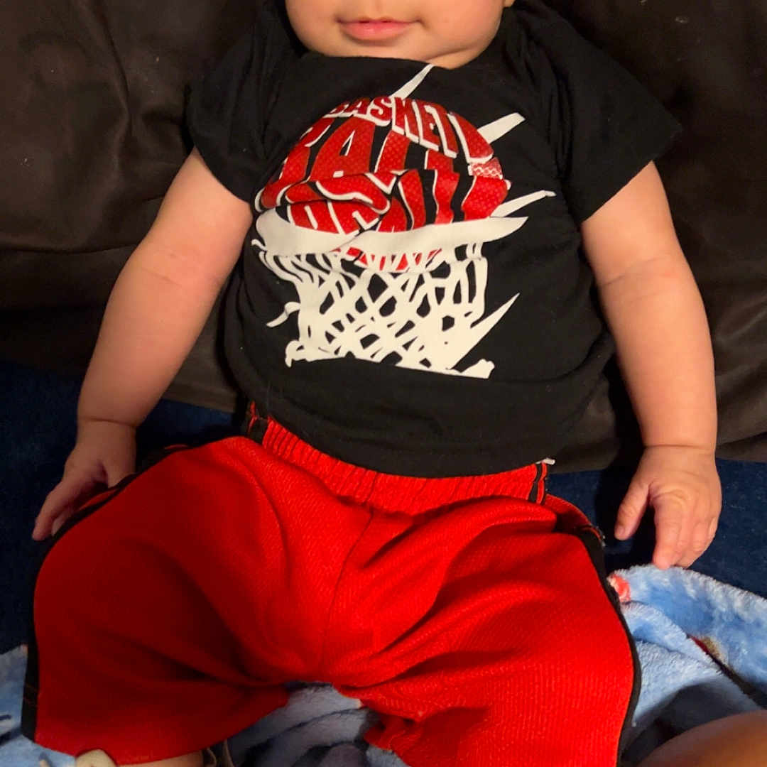 Dominic is registered to the contest to win money with this photo: baby, child, infant, portrait, sitting, smiling, wide_eyed, basketball, sportswear, black_shirt, red_pants, sneakers, shoes, couch, blanket, indoor, home, face, clothing, photograph