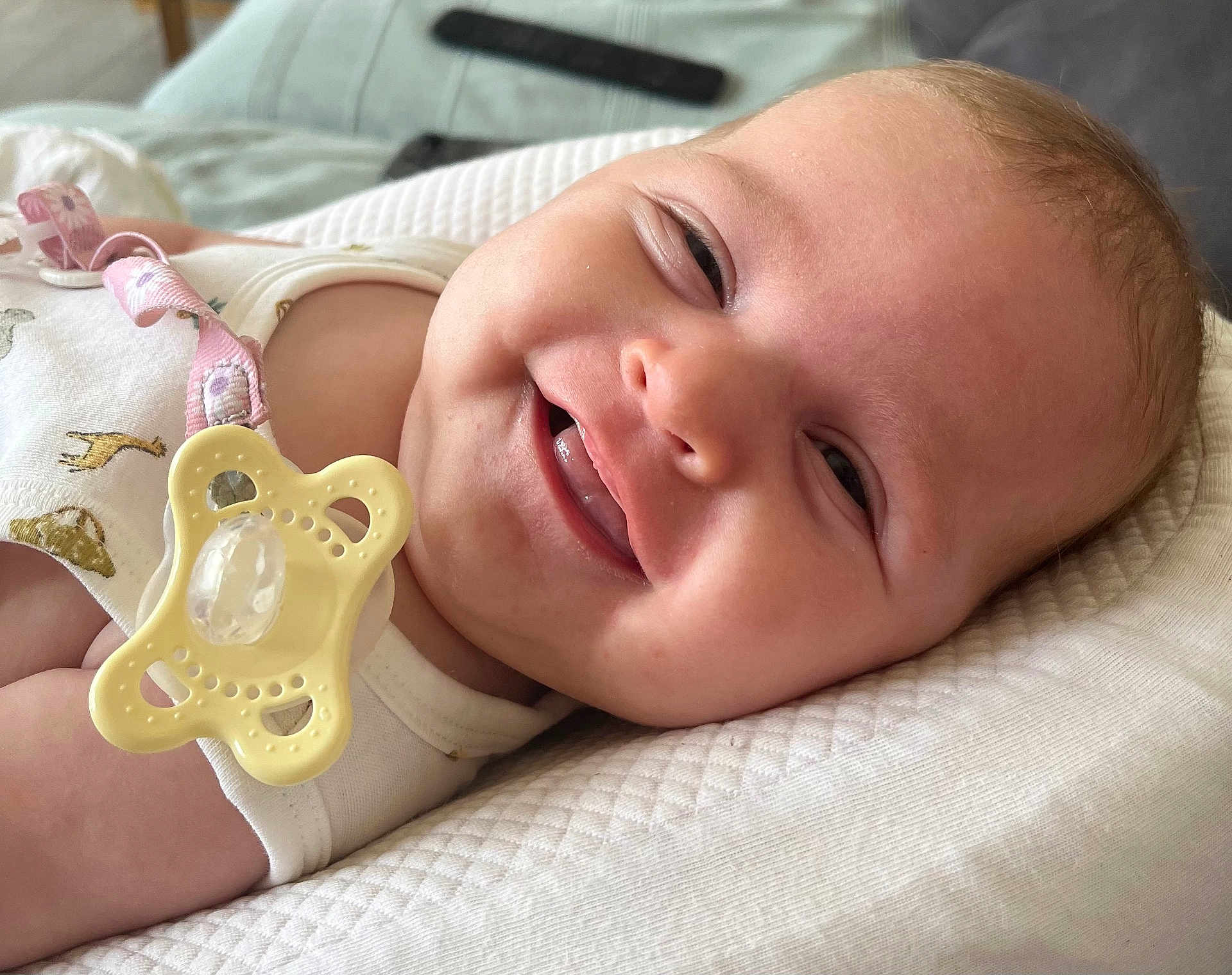 Lou a rejoint le concours — aidez-le/la à gagner de superbes lots ! baby, smiling, pacifier, white_clothing, animal_print, pillow, close_up, infant, cute, happy, skin, face, child, lying_down, indoors, natural_light, texture, relaxed, pacifier_clip, soft