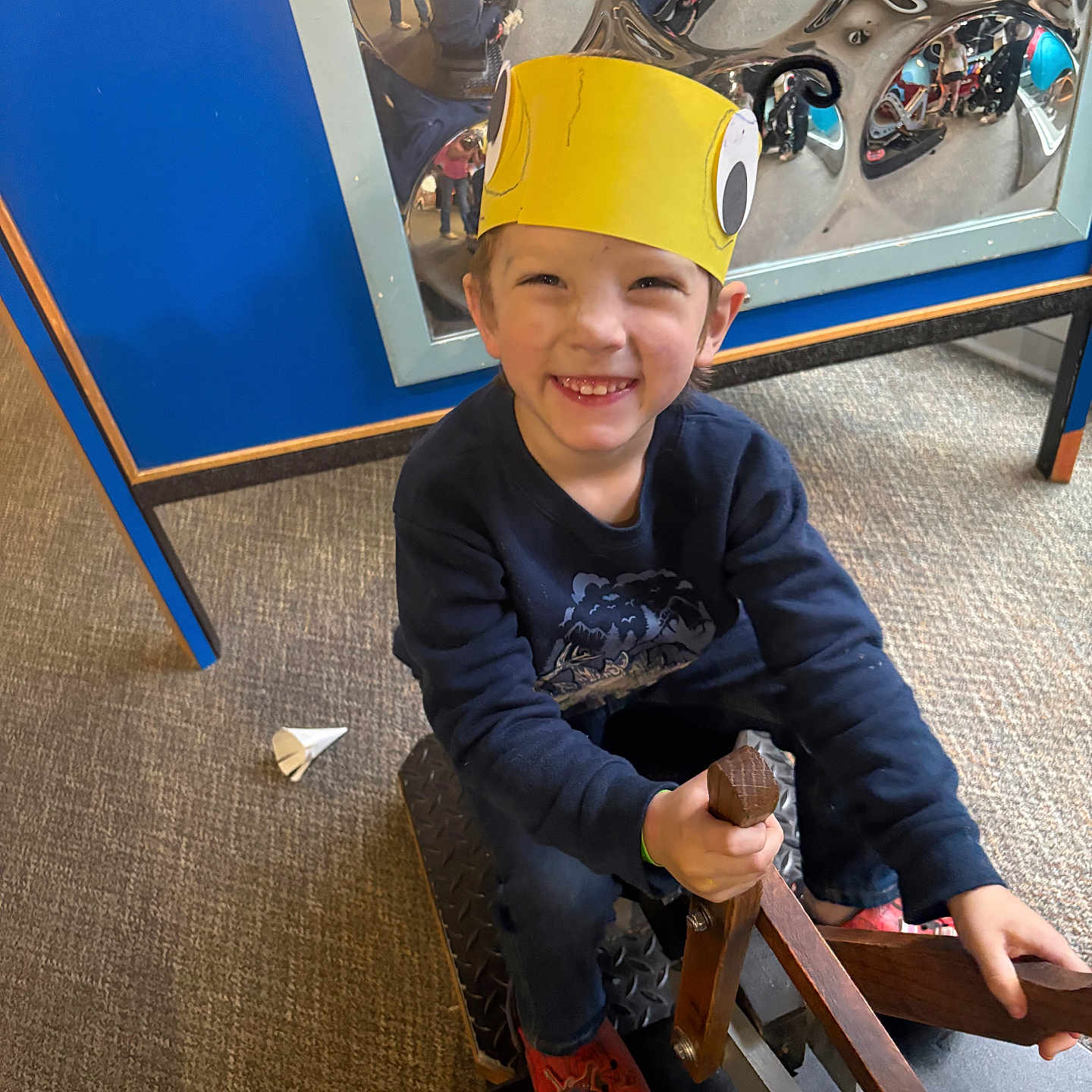 Nathaniel is registered to the contest to win money with this photo: child, boy, smiling, crown, yellow_crown, indoor, play_area, rocking_horse, wooden_toy, reflection, mirror, carpet, casual_clothing, red_shoes, blue_sweater, happy, playful, people, distorted_reflections, toy