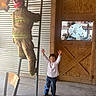 Nathaniel joined the competition — help win amazing prizes! child, firefighter, mannequin, ladder, indoor, wooden_door, corrugated_metal_wall, clothing, jeans, shirt, smiling, person, pose, floor, chair, casual, helmet, reflective_stripes, playful, rustic