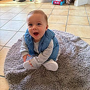 Alessio participe au concours pour gagner de l'argent avec cette photo : baby, child, smiling, indoor, rug, blue_vest, gray_outfit, floor_tiles, vacuum_cleaner, giraffe_plush, toy, happy, sitting, footwear, person, home, playful, young_child, floor, carpet