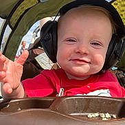 Emory is registered to the contest to win money with this photo: toddler, child, earmuffs, stroller, red_shirt, smile, hand, outdoor, sunlight, face, baby, seat, people, sky, daylight, food, tray, person, blurred_background, casual