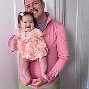 Willow is registered to the contest to win money with this photo: baby, man, child, smile, happy, holding, dress, peach, pink, green_shorts, indoor, person, family, portrait, cute, flower_dress, baby_bow, joy, parent, door