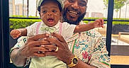 Sire is registered to the contest to win money with this photo: baby, child, man, smile, happy, indoor, window, greenery, table, hat, bowtie, patterned_shirt, beard, watch, hand, holding, portrait, people, family, casual