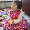 toddler, child, smiling, sitting, toy_block, colorful_dress, quilt, indoor, happy, playtime, barefoot, home, window, furniture, floor, earrings, long_sleeve_shirt, hair, face, person