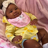 Nyla joined the competition — help win amazing prizes! baby, blanket, bow, cheeks, couch, cute, footwear, hand, happy, headband, indoor, infant, lying_down, pink_bib, relaxing, skin, smile, soft_texture, toddler, yellow_clothing
