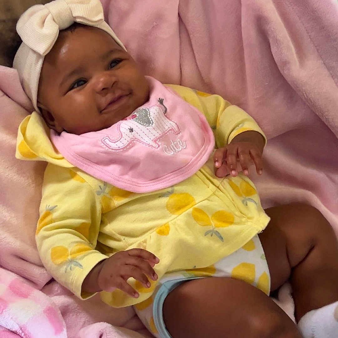 Nyla joined the competition — help win amazing prizes! baby, blanket, bow, cheeks, couch, cute, footwear, hand, happy, headband, indoor, infant, lying_down, pink_bib, relaxing, skin, smile, soft_texture, toddler, yellow_clothing