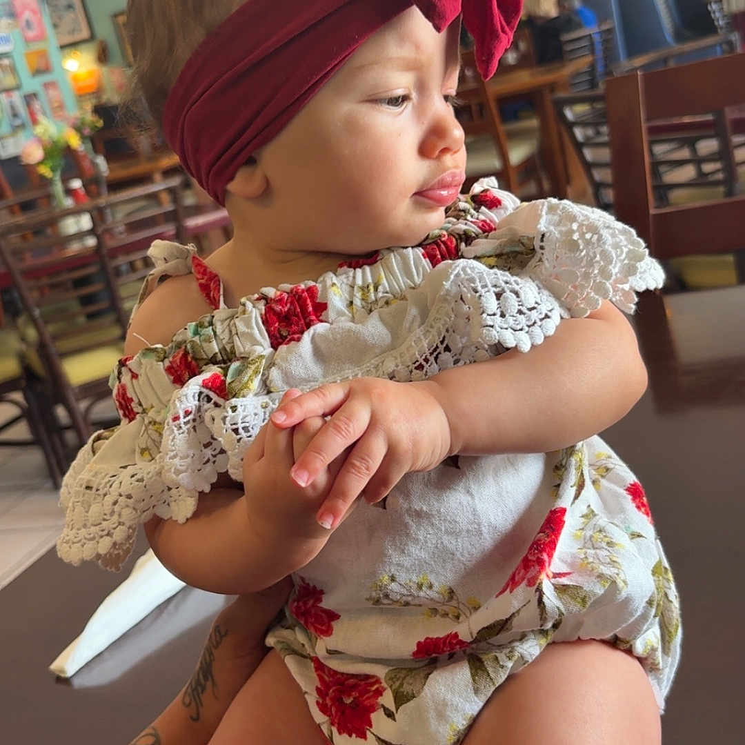 Ariyana is registered to the contest to win money with this photo: arm, baby, bow, cafe, chair, child, clothing, cute, floral_dress, hands, headband, indoor, infant, person, seated, side_view, skin, table, tattoo, toddler
