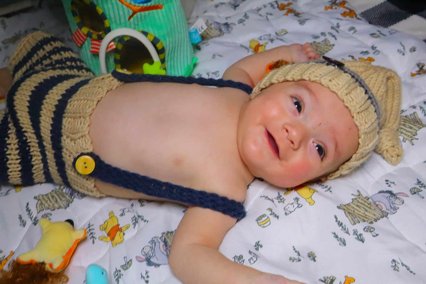 Liam is registered to the contest to win money with this photo: baby, child, knitted_hat, striped_clothing, blanket, toy, smile, lying_down, cute, infant, face, happy, soft_toy, colorful, indoor, playtime, young_child, headwear, portrait, cozy