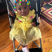 Jream joined the competition — help win amazing prizes! baby, carpet, child, clothing, colorful_beads, curious_expression, cute, footwear, hair_accessories, home, indoor, patterned_rug, person, portrait, seated, socks, stroller, toddler, wooden_floor, yellow_dress