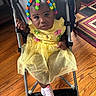 baby, carpet, child, clothing, colorful_beads, curious_expression, cute, footwear, hair_accessories, home, indoor, patterned_rug, person, portrait, seated, socks, stroller, toddler, wooden_floor, yellow_dress